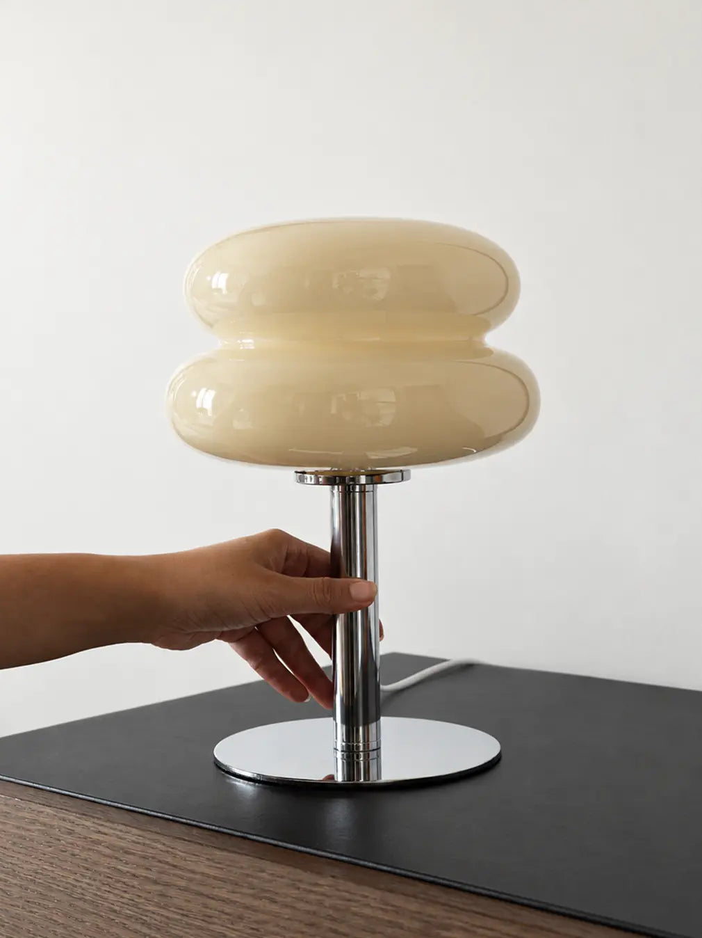 Hand holding the chrome stem of an Orbit yellow glass table lamp on a desk.