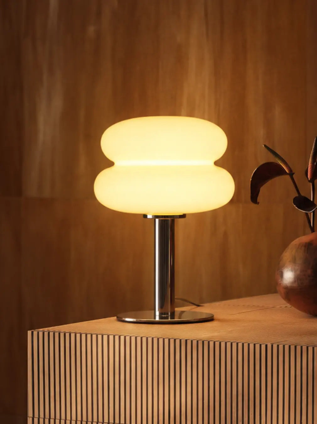 Orbit yellow glass table lamp glowing on a fluted wooden cabinet beside a vase.