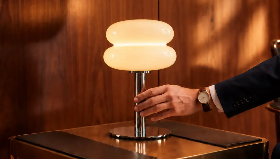 Hand holding the chrome stem of a glowing Orbit yellow glass table lamp on a cabinet.
