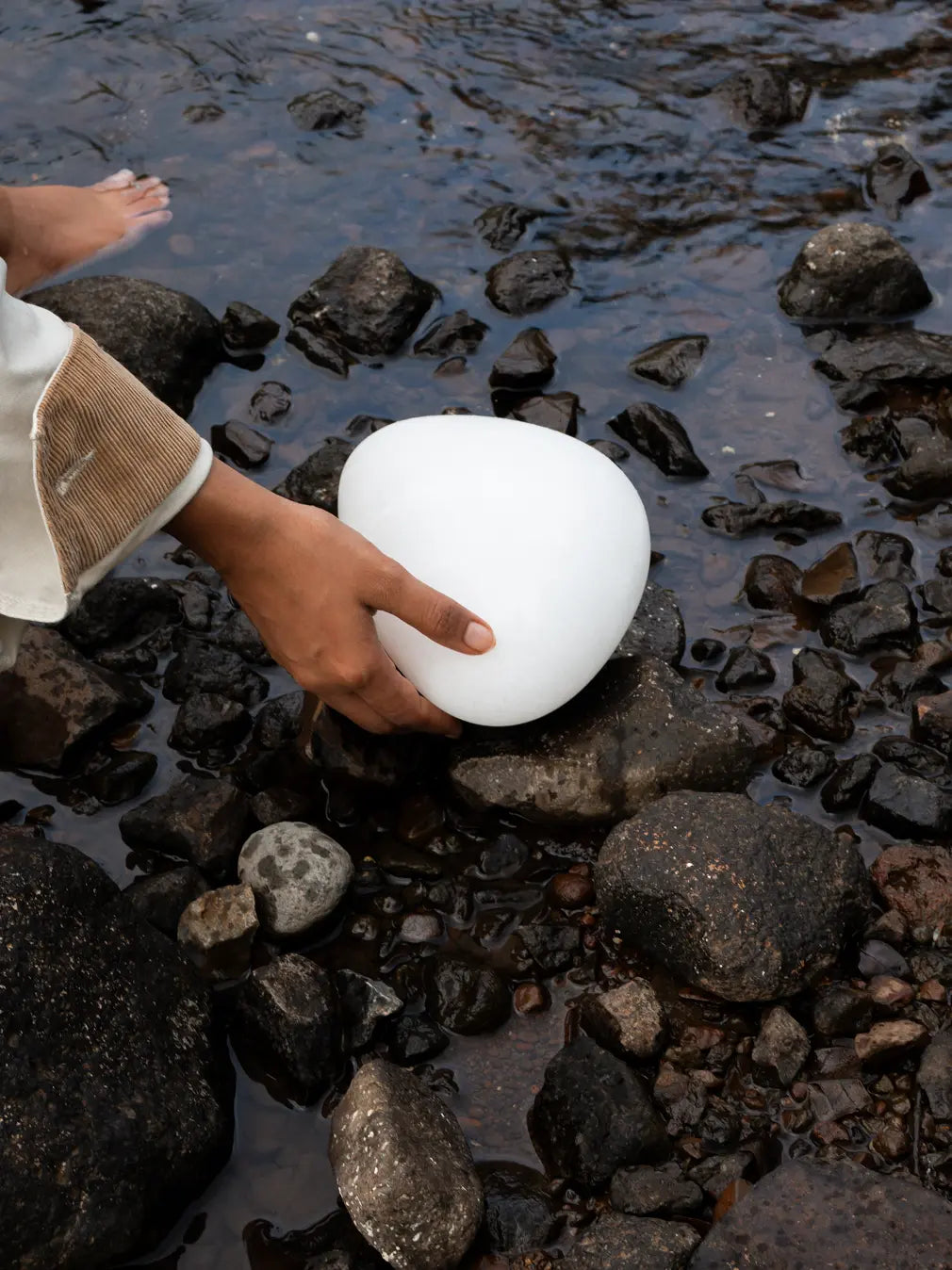 Hand placing the white Pebble Marble Portable Table Lamp on a rock beside a stream